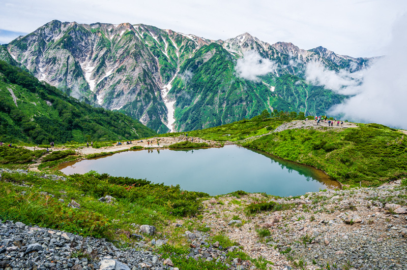 Happo Pond, a small body of water on the ridgeline of Happo, sits below a mountain range with several rocky peaks