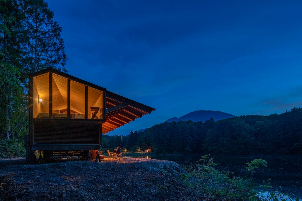 A glamping trailer cabin sits beside the shore of a pond at night. Warm light emits from its large windows.